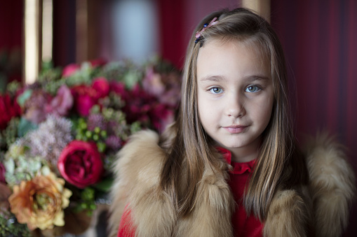 A young green-eyed girl smiles, dressed in a fur waistcoat. In the background, there is a mirror and some flowers