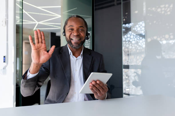 istock Help desk worker inside office at workplace, man smiling and looking at camera, waving greeting gesture, talking remotely, using headset phone and tablet for video calling and consulting customer support 1862684729
