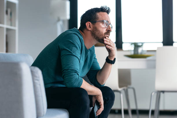 Worried mature man thinking about what he has read on his mobile phone while sitting on the couch at home