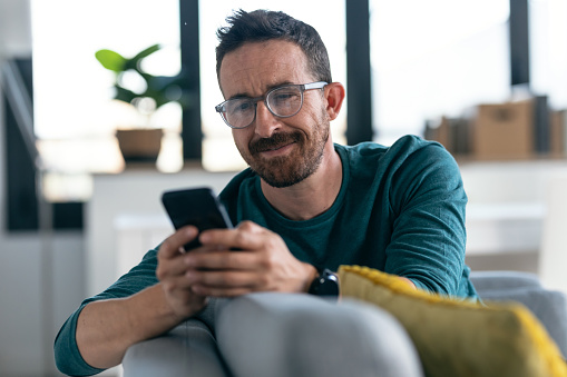 Shot of happy mature man with eyeglasses using his smartphone while relaxing in the couch at home.