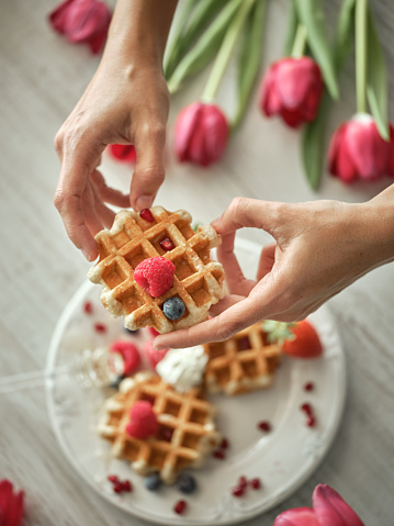 From above crop anonymous person holding tasty homemade waffle with berries while standing at table with tulips in light kitchen