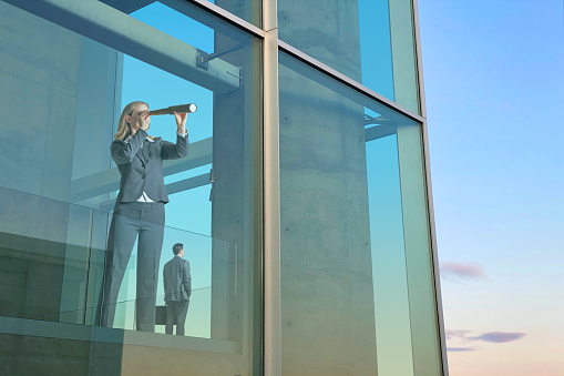 A businesswoman stands at a large office building window as she looks out through a spyglass.  A male business colleague looks out another window as he stands behind her.
