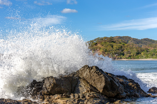 Nature landscape showing a powerful wave crashing into a rock in Costa Rica.  A forested island is seen in the background.  Concepts could include nature, travel, power, other.