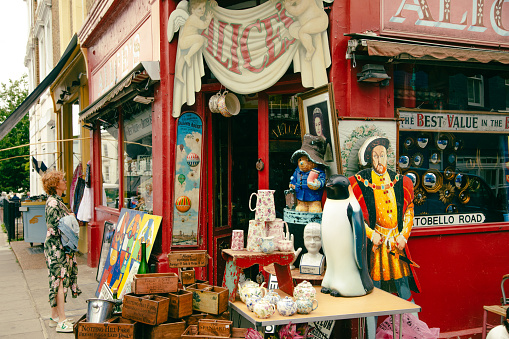 A glimpse into the vibrant world of Portobello Road in London, where people are captivated by the charm of Alice's Antiques. Passersby stop to peer through the window display, drawn by the allure of unique treasures within. The eclectic mix of antiques tells stories of a bygone era, inviting curiosity and sparking the imagination. Portobello Road, London, antique shop, window shopping, vintage treasures, eclectic collection, antiques market, curiosity, historical artifacts, unique finds, exploring, street scene, cultural heritage, collectibles, hidden gems, city life, passerby, urban culture, London attractions, travel, British traditions