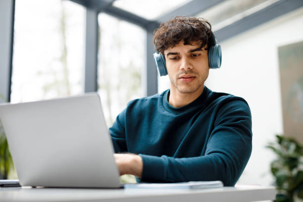 istock Focused guy student in wireless headphones using laptop and checking information in workbook, watching online tutorial 1862898233