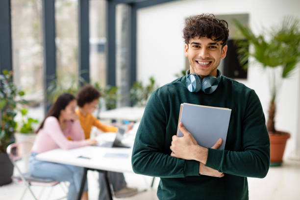 istock European man student holding books and smiling at camera while female classmates studying with laptop on the background 1862898243