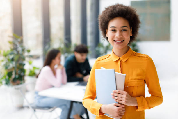 istock Portrait of happy black lady university student posing with notepads while classmates studying on background in audience 1862898799