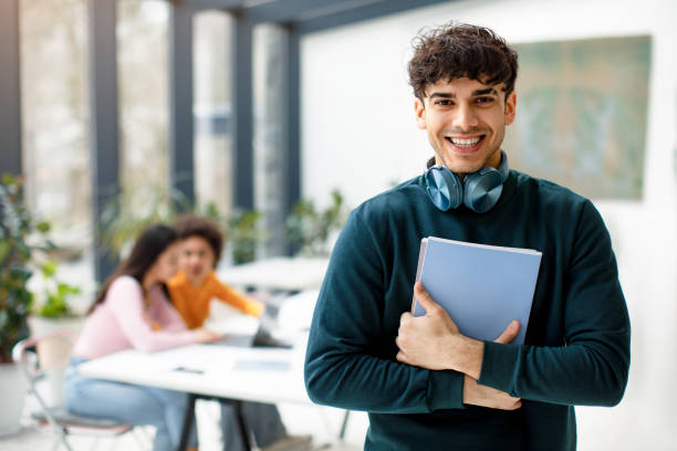 istock Happy student guy posing with notepads in hands and headphones on neck, smiling at camera while classmates studying 1862899340