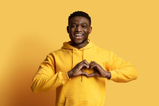 Cheerful handsome millennial black guy wearing yellow hoodie holding hands on chest in heart-shaped symbol and smiling, expressing love isolated on yellow background, copy space
