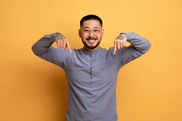 Look At This. Smiling Asian Guy Pointing Down With Two Hands Look At This. Smiling Asian Guy Pointing Down With Two Hands, Cheerful Millennial Man Demonstrating Free Copy Space For Your Design And Advertisement While Standing Over Yellow Background man hand down stock pictures, royalty-free photos & images