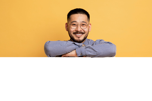 Happy Young Asian Man Leaning At Blank White Advertisement Board, Smiling Millennial Man In Eyeglasses Demonstrating Free Copy Space For Your Ad Design, Standing Over Yellow Studio Background