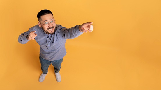 Check This. Handsome asian guy pointing away with two fingers, smiling millennial male in eyeglasses indicating copy space while standing on yellow studio background, above view, panorama