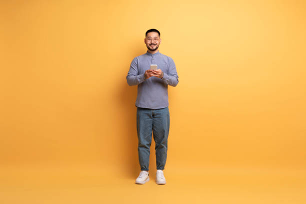 istock Great App. Smiling asian man with smartphone standing on yellow background 1862957778