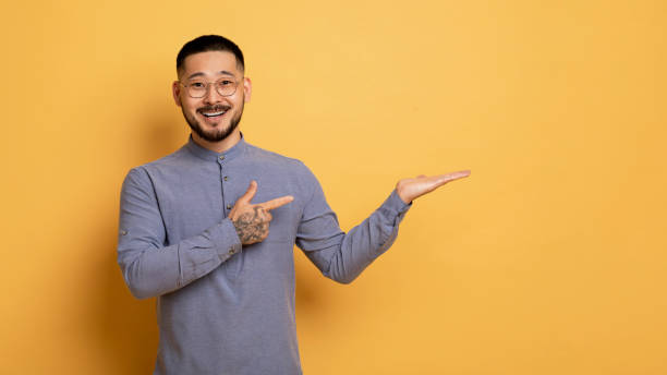 istock Cheerful Asian Guy Pointing Finger At Open Palm, Standing On Yellow Background 1862958291