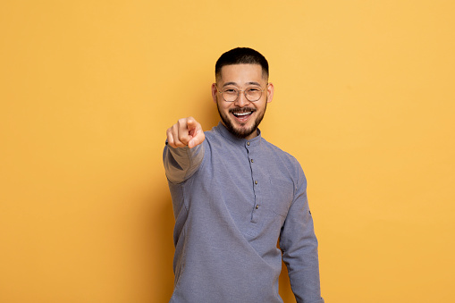 Portrait of young asian man pointing index finger at camera, cheerful millennial guy posing over yellow studio background, smiling male picking, choosing and indicating somebody, copy space