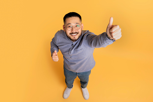Above Shot Of Cheerful Asian Man Showing Thumbs Up At Camera, Happy Excited Millennial Male Gesturing Sign Of Approval While Standing Over Yellow Studio Background, Top View, Copy Space