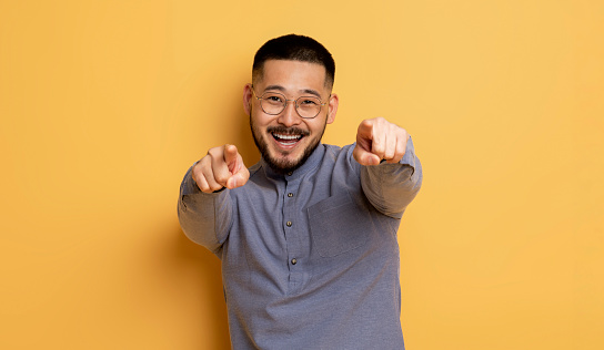 Gotcha. Positive Young Asian Guy Pointing Two Fingers At Camera, Portrait Of Cheerful Millennial Man In Eyeglasses Indicating Somebody While Standing Isolated On Yellow Background, Copy Space