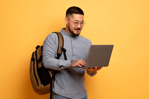 istock Young Asian Student Guy With Laptop And Backpack Standing On Yellow Background 1862958335