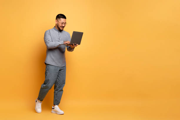 istock Cheerful Young Asian Man With Laptop Walking Over Yellow Studio Background 1862958340