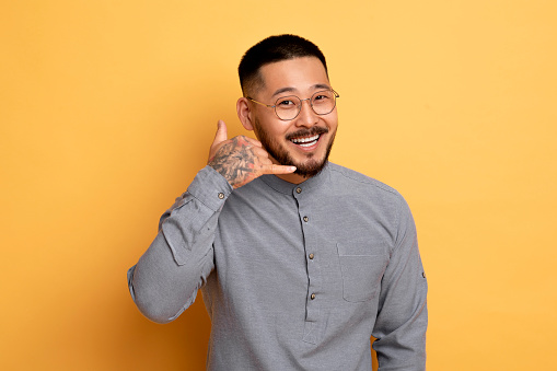 Call Me. Cheerful Young Asian Man With Hand Near Ear Imitating Phone And Looking At Camera, Positive Millennial Guy Wearing Eyeglasses Inviting For Communication While Standing On Yellow Background