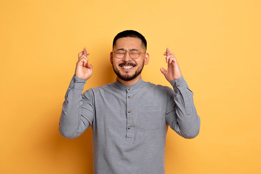 Making Wish. Portrait Of Cheerful Young Asian Man Keeping His Fingers Crossed, Superstitious Millennial Guy Hoping For Luck While Standing With Closed Eyes Over Yellow Studio Background, Copy Space