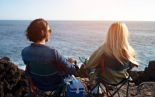 Rear View Of Young Couple Relaxing In Camping Chairs On Beach Rocks Near Ocean, Man And Woman Looking At Sky And Sunset View, Enjoying Outdoor Leisure, Having Picnic Near Water, Free Space