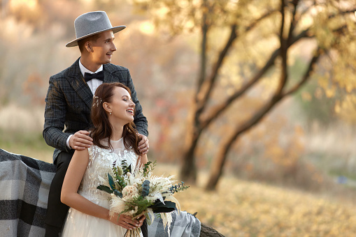 beautiful happy stylish bride and groom in hat near tree in autumn