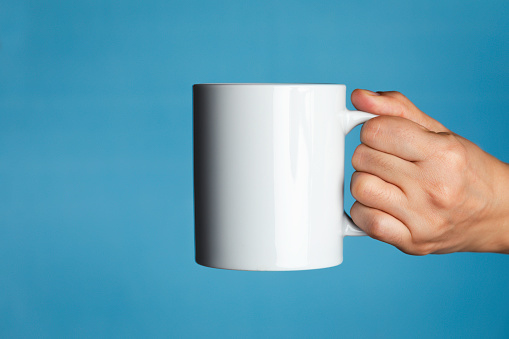 Human hand is holding a white coffee mug  in front of a blue background.