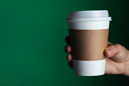 Hand is holding a disposable cup in hand in front of dark green background.