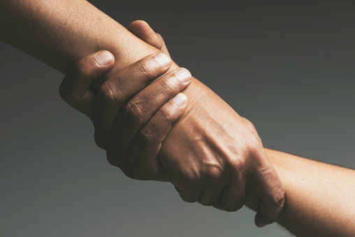 Two unrecognizable caucasian peoples hands are holding each others wrist in front of dark gray background. Arms are forming a diagonal line from the left upper corner to the right bottom corner.
