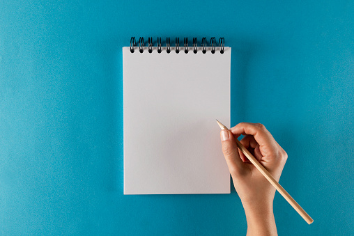 Hand is holding pencil and is about to write something into a spiral notepad on blue background.