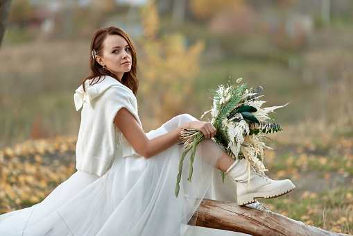 beautiful bride wearing white boots sitting on tree and holding autumn bouquet.