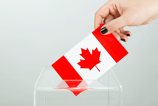 Human hand with black nail polish is inserting flag of Canada into ballot box.