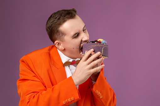 birthday man in orange jacket bites cake on purple background. copy space