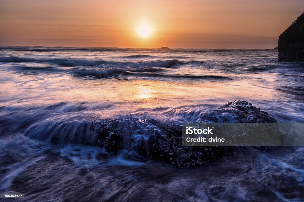 Amethyst Tide Ocean tides crash over rocks  forming amethyst colored waters against a soft sunset in this sunset tides background image. Horizontal Stock Photo
