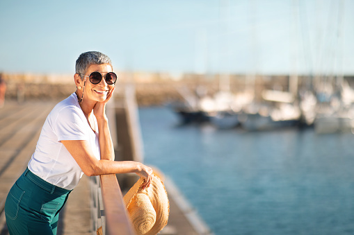 Enjoy Golden Summer. Cheerful European Senior Woman Standing At Marina Pier, Posing With Sunglasses And Straw Hat Outdoors, Enjoying Dream Vacation By The Sea. Copy Space