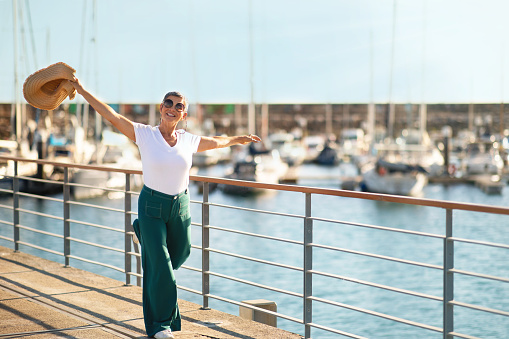 Golden Summer Holidays. Joyful Gray European Lady Posing With Straw Hat And Having Fun At Marina Dock Outside, Near Parked Luxury Yachts, Celebrating Vacation And Retirement Freedom. Copy Space