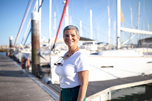 Sailboat Sailing. Happy Mature Woman With Short Haircut Standing Near Yachts At Marina Pier, Waiting For Luxury Sea Cruise. Lady Enjoying Dream Summer Vacation, Posing Looking Aside Outdoor
