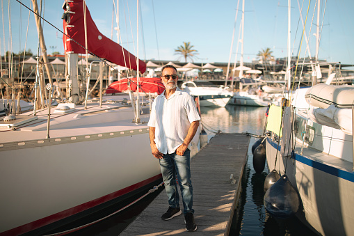 Yacht Owner. Tanned Mature Man On Vacation Posing Near Luxury Sailboat Outdoor, Standing With Confidence At Marina Boats Park Outside, Full Lengt Shot. Summer Tourism And Seaside Life Concept