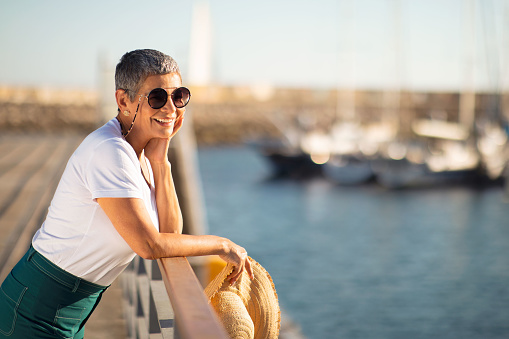 Coastal Relax. Delighted senior woman wearing her sunglasses and holding beach hat at the marina, looking at sailboats. Tourist embracing sea breeze and holiday spirit by boats, free space