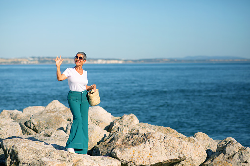 Hello, Summer. Happy European Mature Lady Waving Hand To Camera Standing Near Sea Shore Outdoor. Woman Enjoying Holidays And Sunny Day At Seaside. Dream Vacation Concept. Full Length