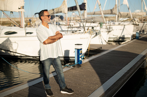 Dream Sea Holidays. Mature gentleman with hands crossed, gazes into distance standing at marina outdoor, surrounded by luxury boats. Seasoned sailor. Maritime experience, vacation. Copy space