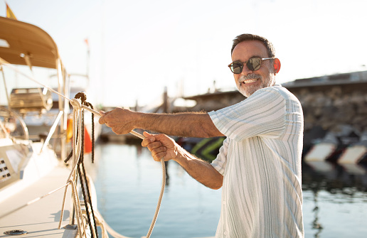 Harbor Skills. Senior man confidently manages the rope, docking his yacht at marina, smiling to camera while demonstrating years of sailing expertise at sunlit pier. Seaside vacation leisure