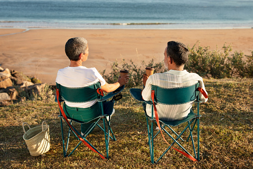 Summer vacation moments. Back view of older couple sitting together facing the ocean and waves, enjoying coffee and taking pause at seaside outdoor. Golden holidays concept