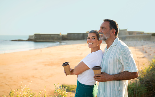 Holidays moments. Mature man wrapping his arms around his wife standing by ocean, both holding paper coffee mugs. Romantic senior couple having perfect summer vacation by coastline. Space for text
