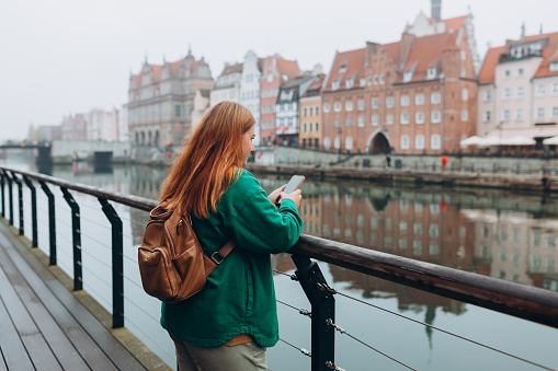 Attractive young female tourist is exploring new city. Redhead girl holding a mobile phone on city street in Gdansk. Traveling Europe in autumn. Vacation concept