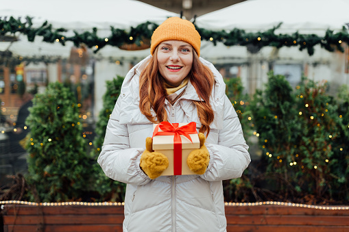 Happy young beautiful excited woman holding gift box in hands and smile on winter street. Winter holidays sale. Concept of shopping, holidays and happiness christmas.