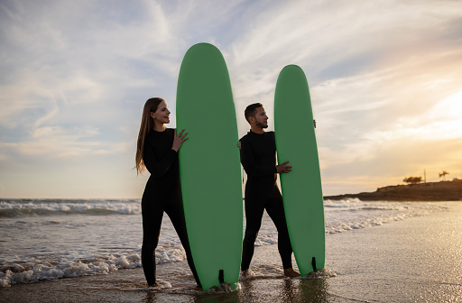 Happy Young Couple In Wetsuits Surfing On The Beach Together, Portrait Of Millennial Surfers Man And Woman Posing With Surfboards In Hands On Ocean Shore, Enjoying Active Lifestyle, Copy Space
