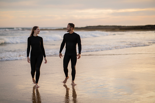 Happy young man and woman in wetsuits walking on the beach at sunset time, romantic millennial couple chilling at seashore, going out of water after surfing together, full length, copy space