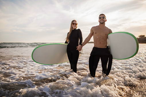 Xtreme Sports. Young Sporty Couple Surfing Together On The Beach, Portrait Of Smiling Man And Woman Wearing Wetsuits Holding Hands And Carrying Surfboards, Millennial Surfers Standing In Water